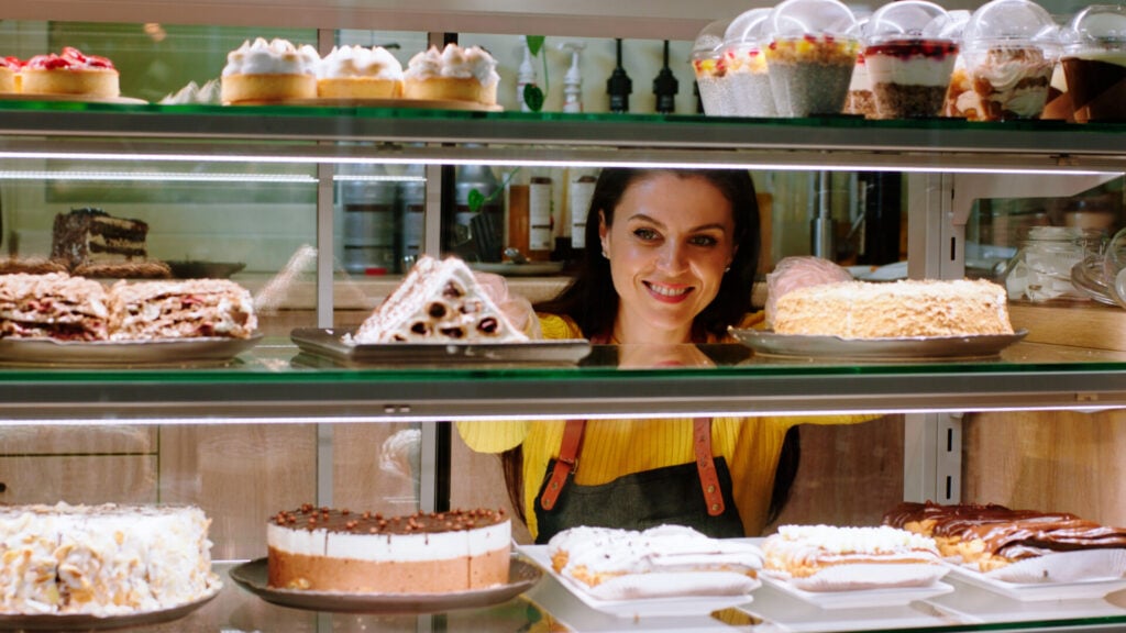 A woman looking at shelves of cakes in a bakery while smiling.